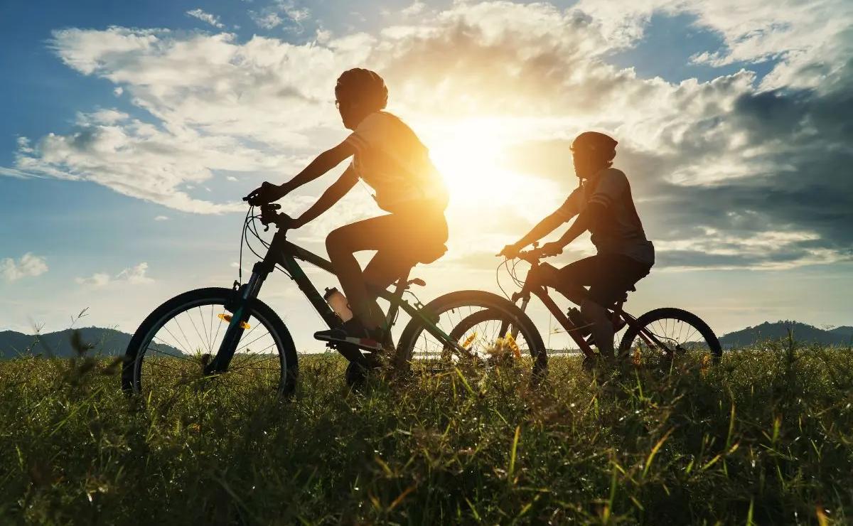 Two people ride bicycles on a grassy field at sunset with the sun and clouds visible in the background.