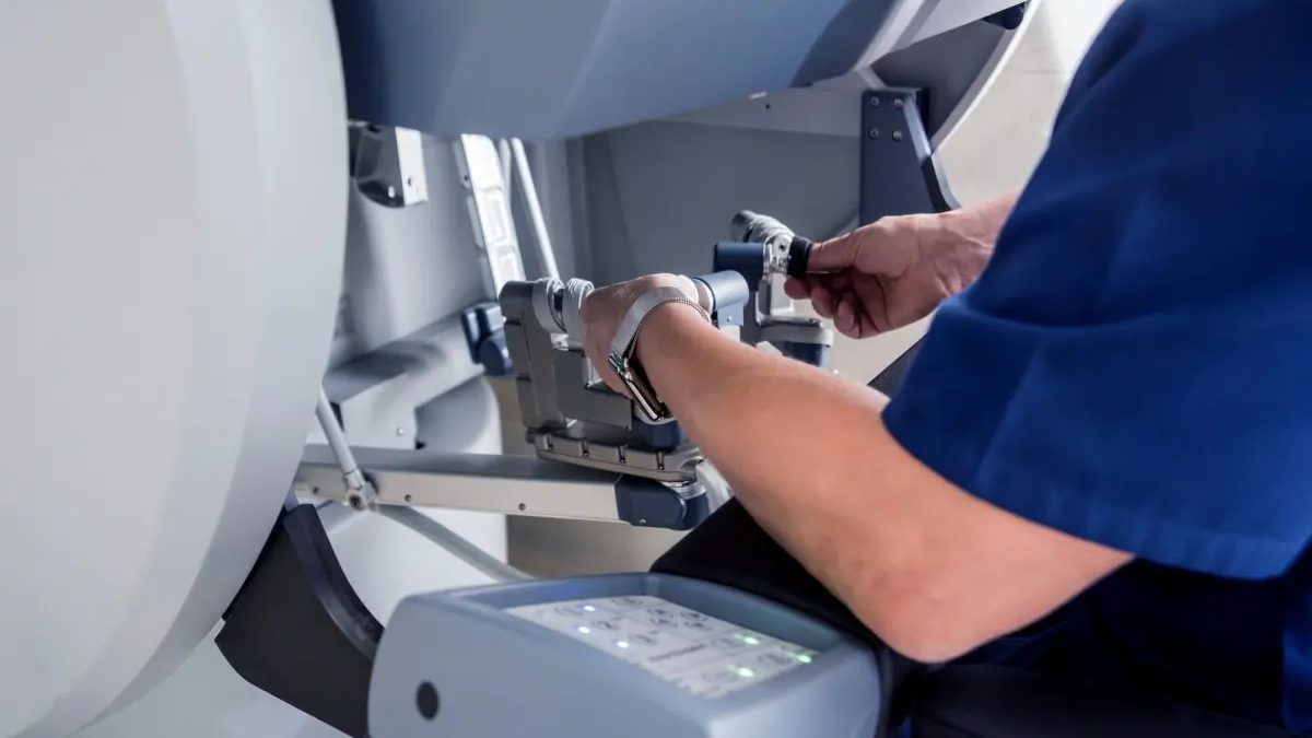 A person in scrubs operates the controls of a robotic surgical system, manipulating handles and buttons on a medical device.