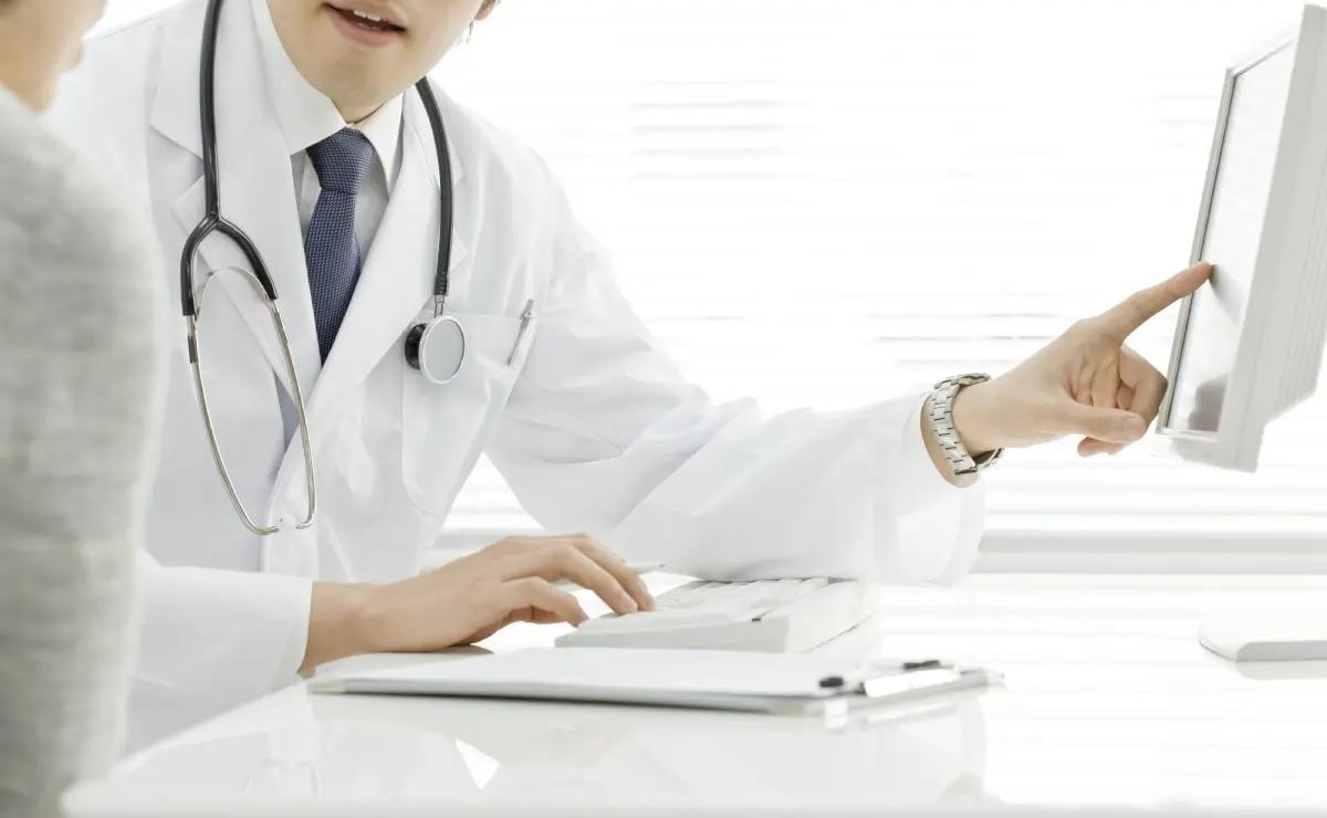 A doctor wearing a white coat and stethoscope points at a computer screen while speaking with a patient.