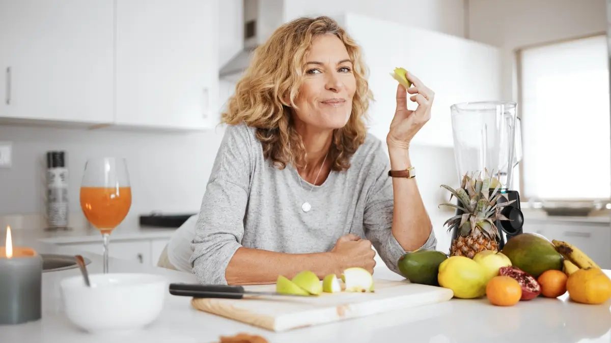 A woman in a kitchen eats a piece of green apple, surrounded by various fruits, a blender, and a glass of juice on the counter.