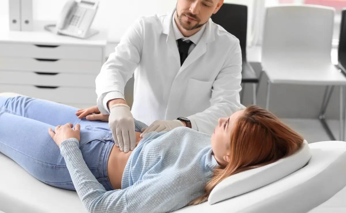 A doctor wearing gloves examines a woman’s abdomen as she lies on an exam table in a medical office.