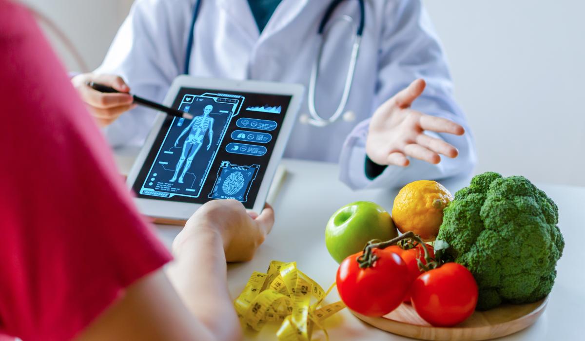 A doctor discusses a digital X-ray with a patient; a plate with vegetables, fruits, and a measuring tape are on the table, suggesting a focus on nutrition and health.