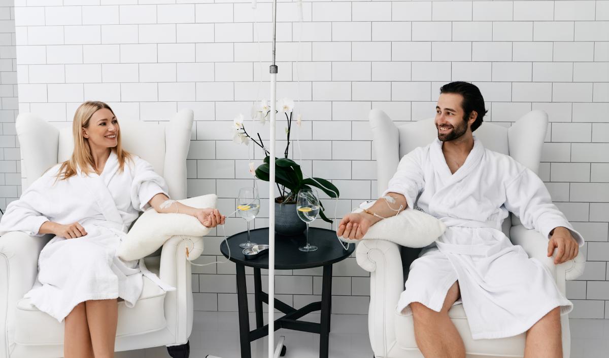 Two people in white robes sit in armchairs receiving IV drips, with a table holding a potted orchid and glasses between them, in a white tiled room.