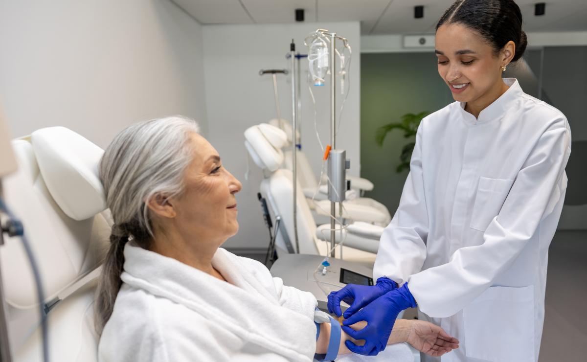A healthcare professional in a white coat and blue gloves prepares to take blood from an older woman seated in a medical clinic.