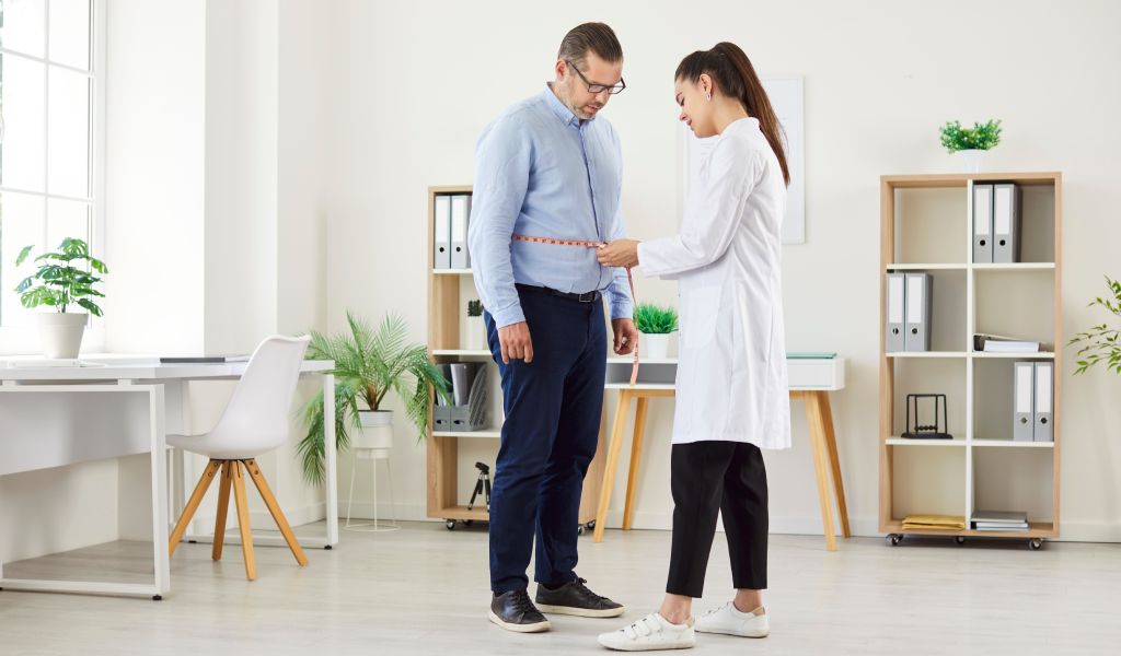 Young female nurse measure waist with measuring tape for mature man patient standing in medical clinic during examination. 