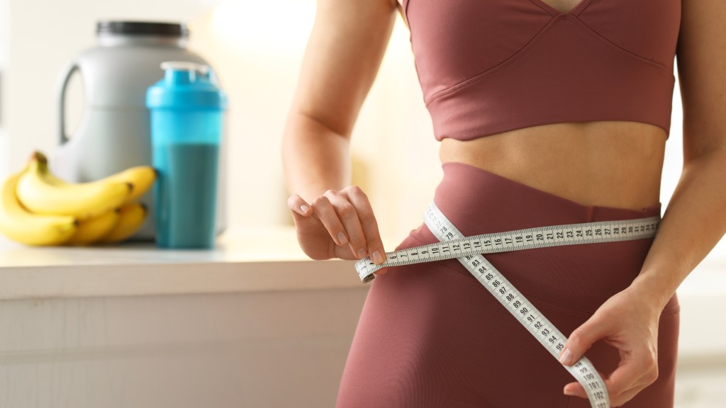 A woman measures her waist in the kitchen, with bananas and a shake in the background – demonstrating the effects of weight loss and healthy eating habits.