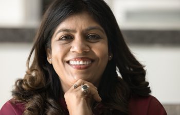 Close facial portrait of happy mature Indian woman with wavy black hair looking at camera, smiling, showing healthy white teeth, touching face, chin, promoting dental care. Cropped head shot, closeup